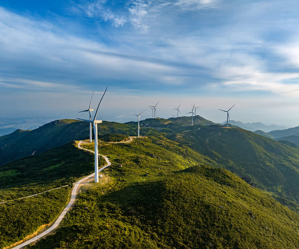 Wind turbines lined along a winding road on green mountainous terrain under a partly cloudy blue sky.