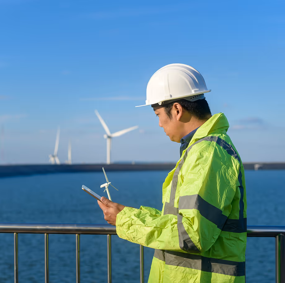 Engineer wearing a white hard hat and neon yellow safety jacket using a tablet near offshore wind turbines over the sea.