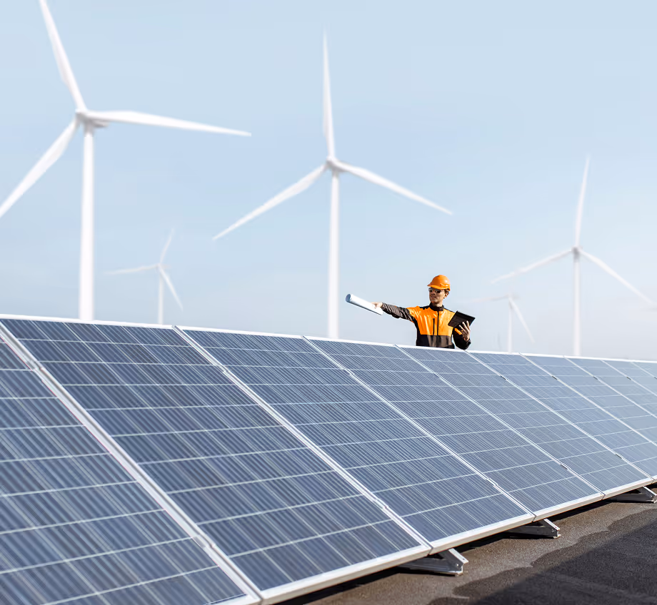 Engineer in orange safety gear holding a tablet and blueprint standing near solar panels with wind turbines in the background under a clear sky.