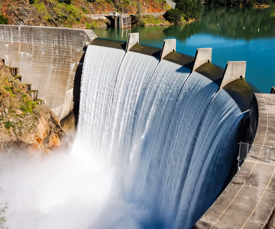 Curved concrete dam spilling water into misty area below, surrounded by rocky hills and calm reservoir.