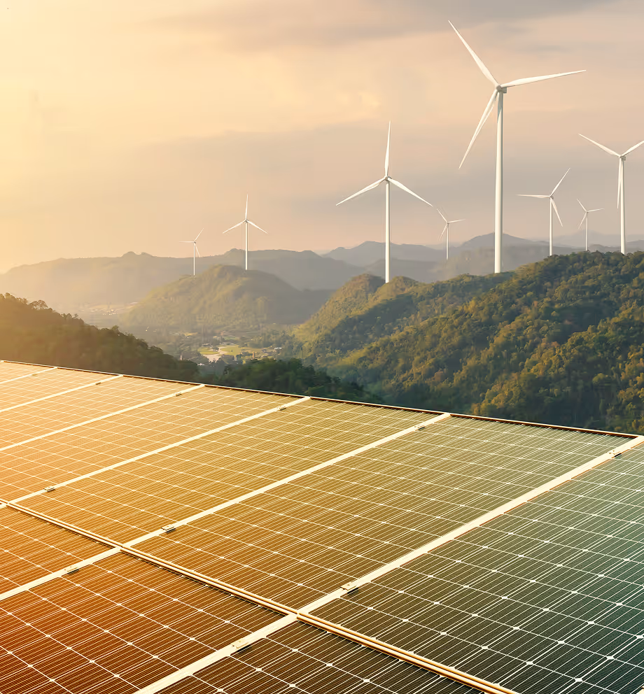 Solar panels in the foreground with wind turbines on green hills under a warm, partly cloudy sky.
