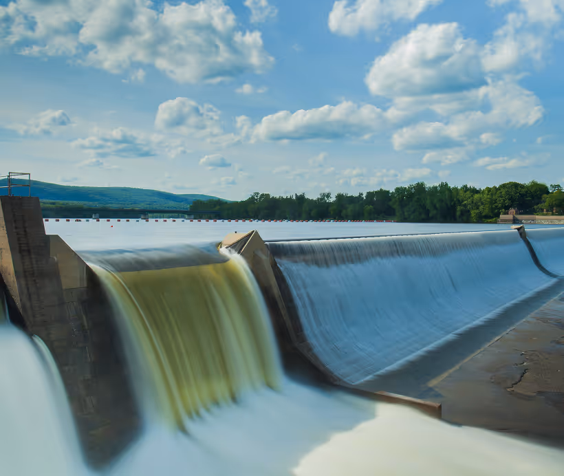 Wide dam with water flowing over the spillway under a blue sky with scattered clouds.