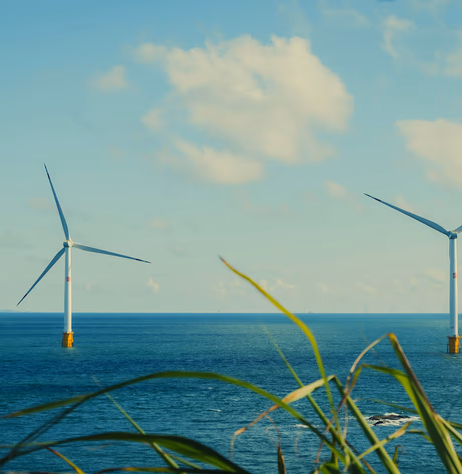 Two offshore wind turbines standing in the ocean with blue sky and some clouds, green grass in the foreground.