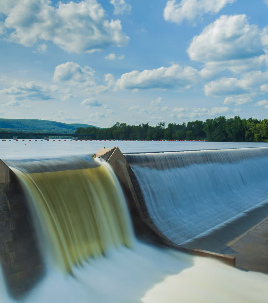 Water flowing over a concrete dam under a blue sky with scattered clouds, surrounded by trees and hills in the background.