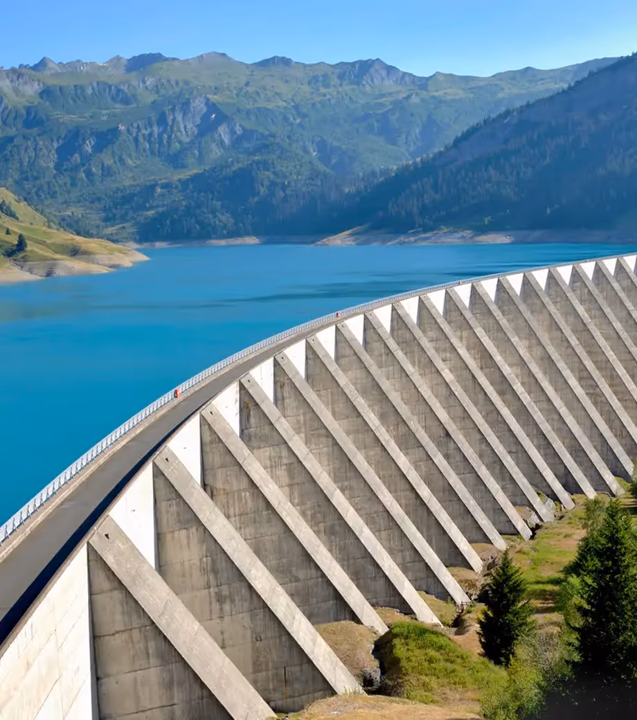 Curved concrete dam holding bright blue water with green mountains and trees in the background under a clear sky.