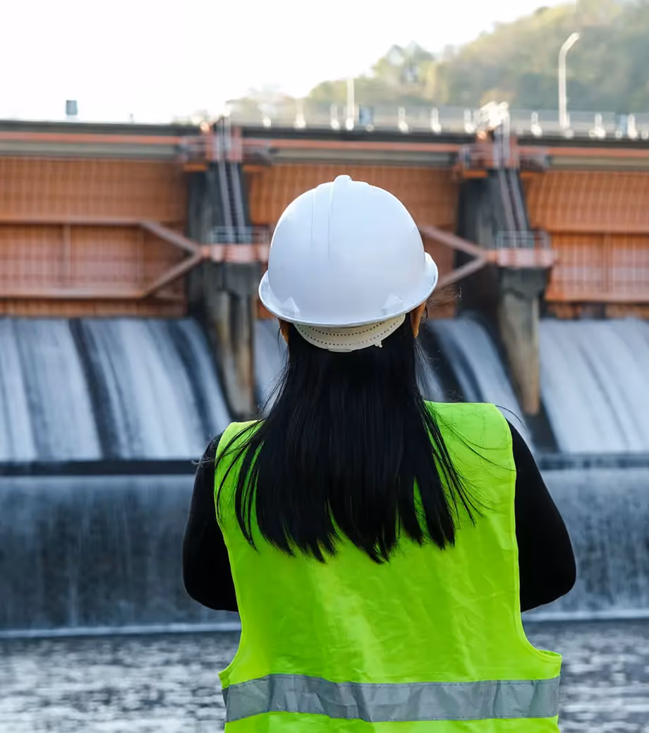Engineer wearing white hard hat and neon green safety vest overlooking a dam with flowing water.