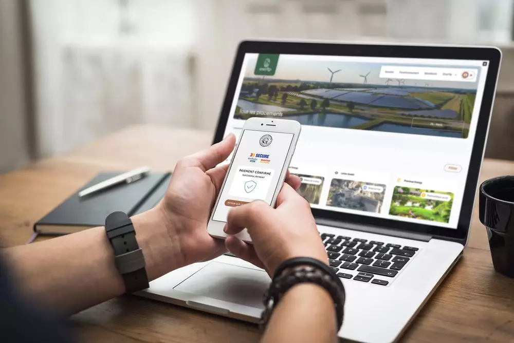 Person holding a smartphone showing payment confirmation screen in front of a laptop displaying renewable energy website with solar panels and wind turbines.
