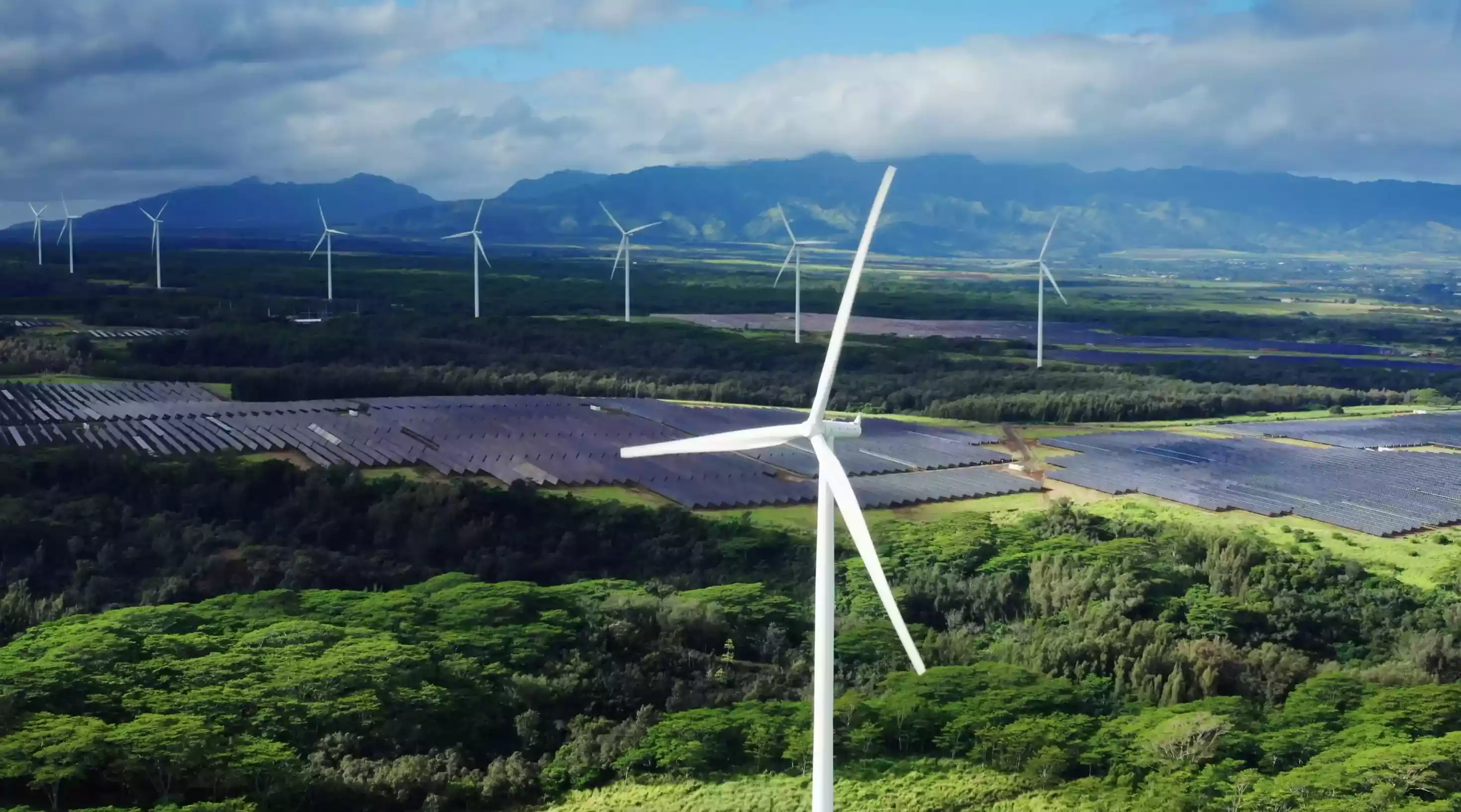 Wind turbines and solar panels in a green landscape with mountains and partly cloudy sky in the background.