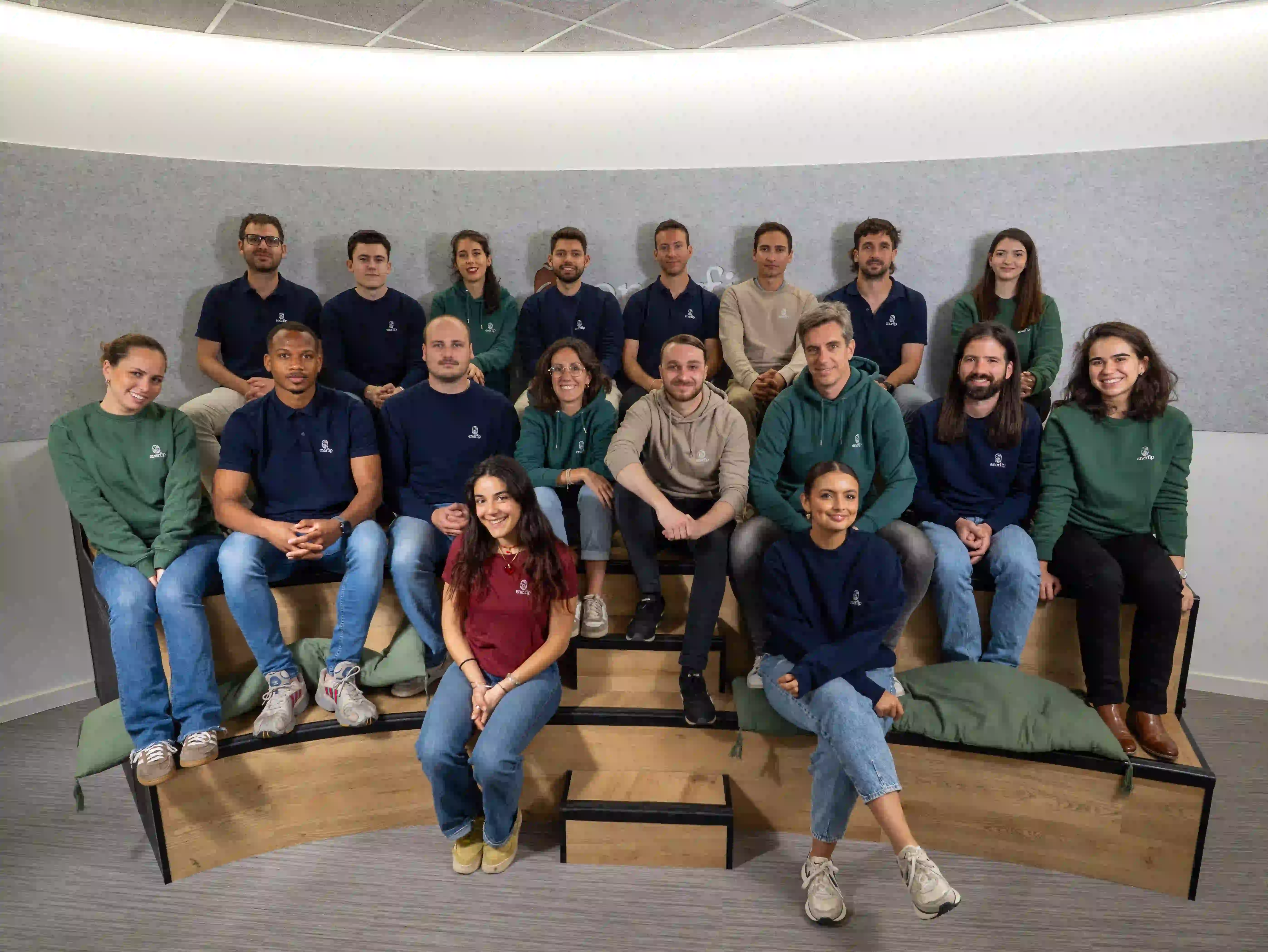 Group of 18 people wearing casual attire with company logos seated on tiered wooden benches in a modern office space.