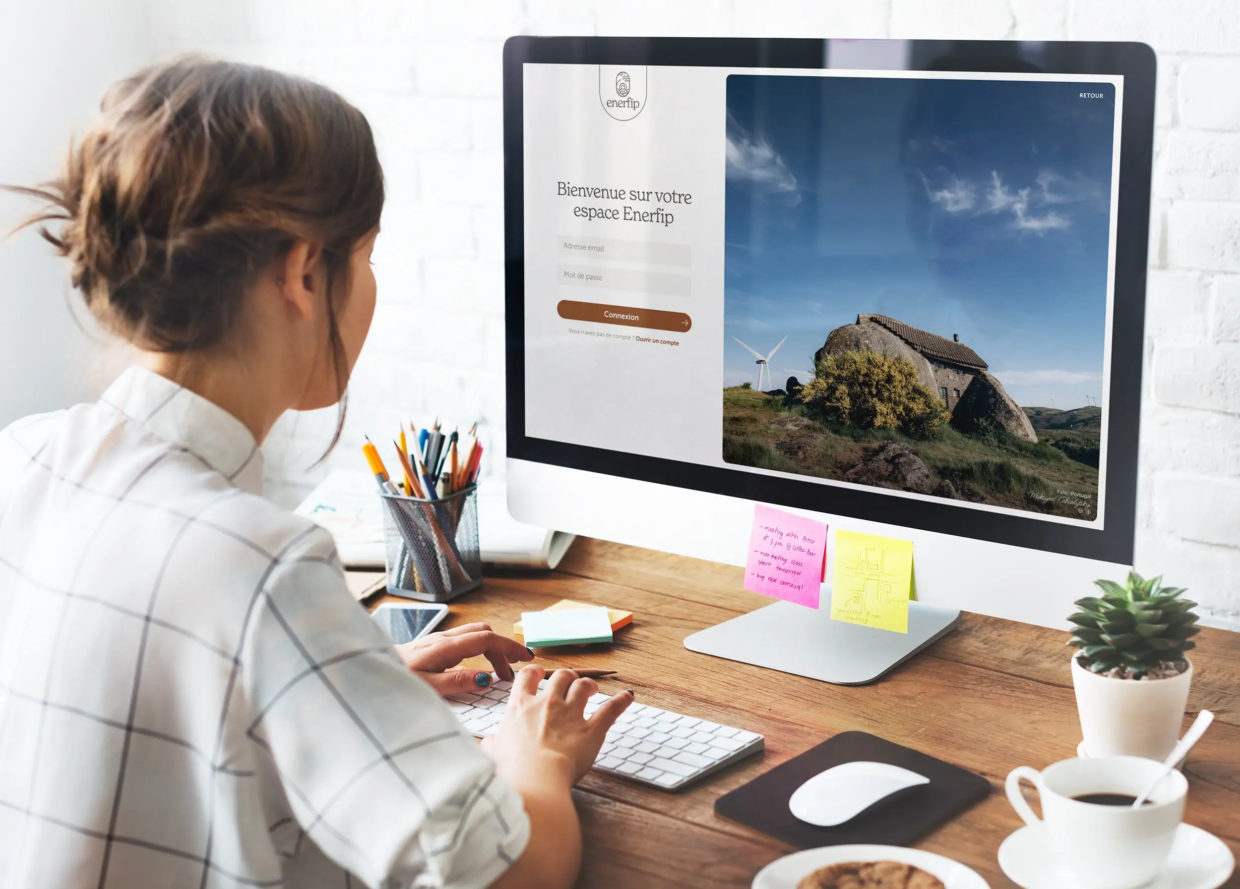Woman with brown hair tied in a bun typing on a keyboard at a desk with a computer screen displaying a French login page and a landscape image.