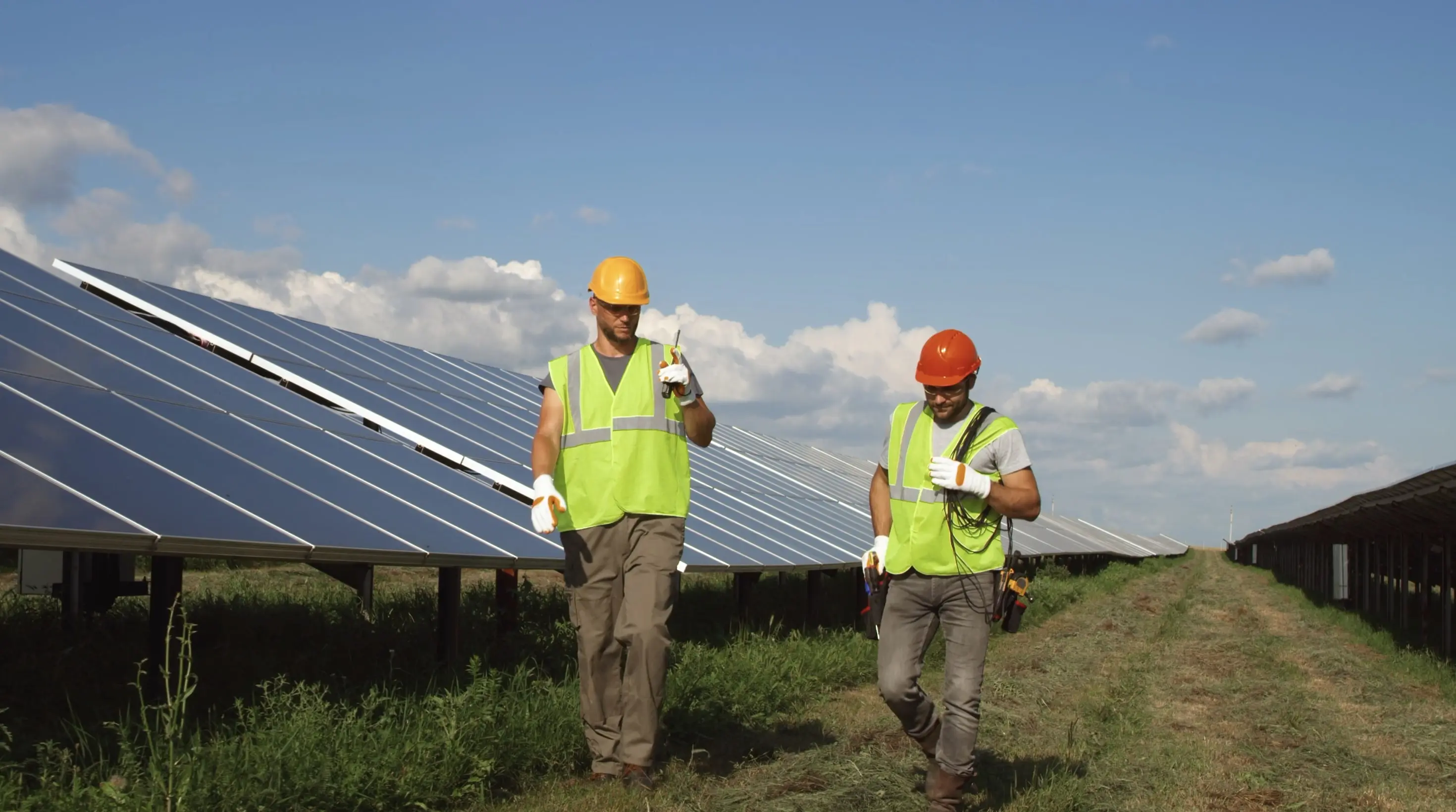 Two workers wearing safety vests and helmets walking beside rows of solar panels under a partly cloudy sky.