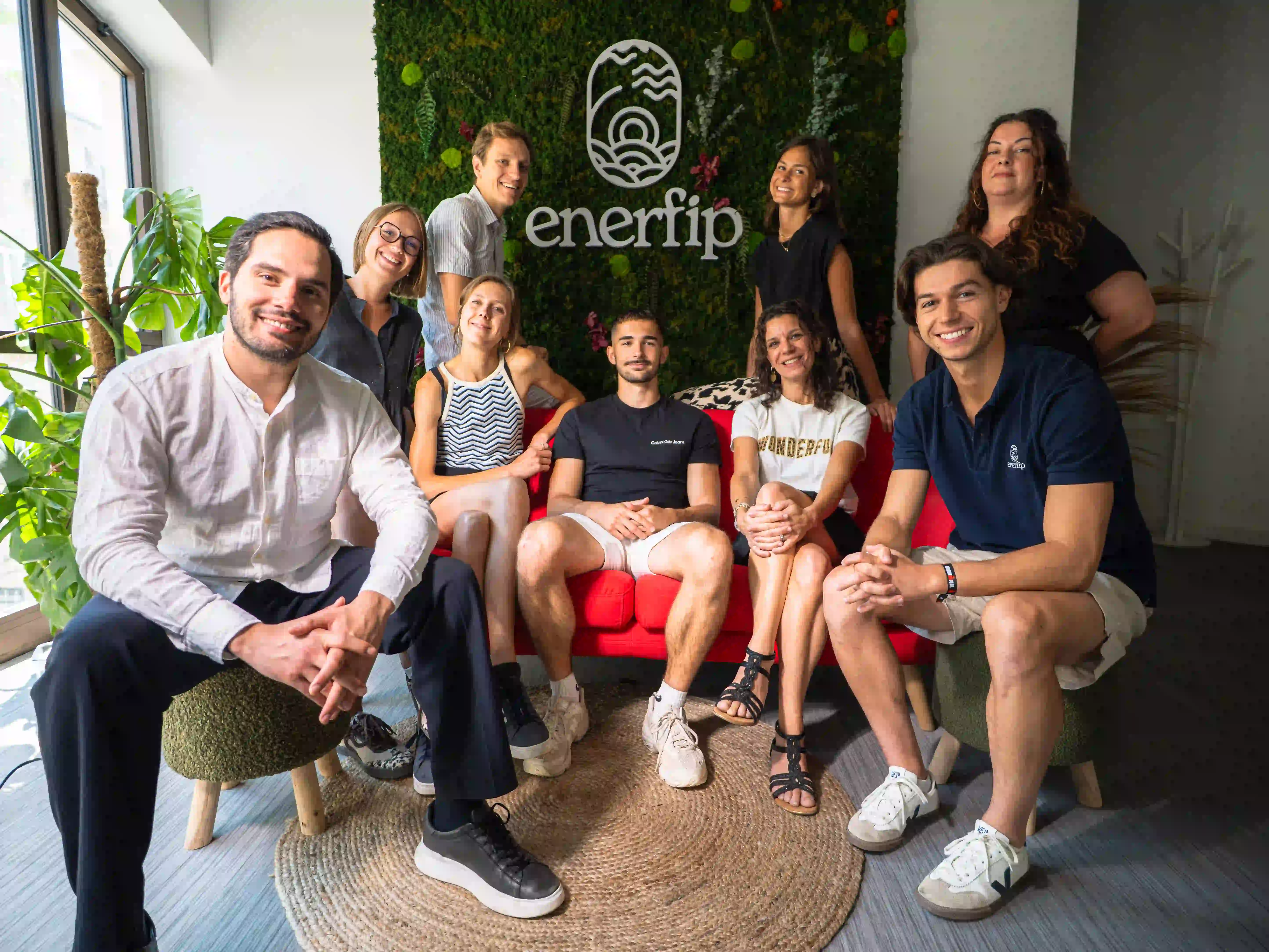 Group of nine young adults smiling and sitting in a modern office space with the Enerfip logo on a green plant wall behind them.