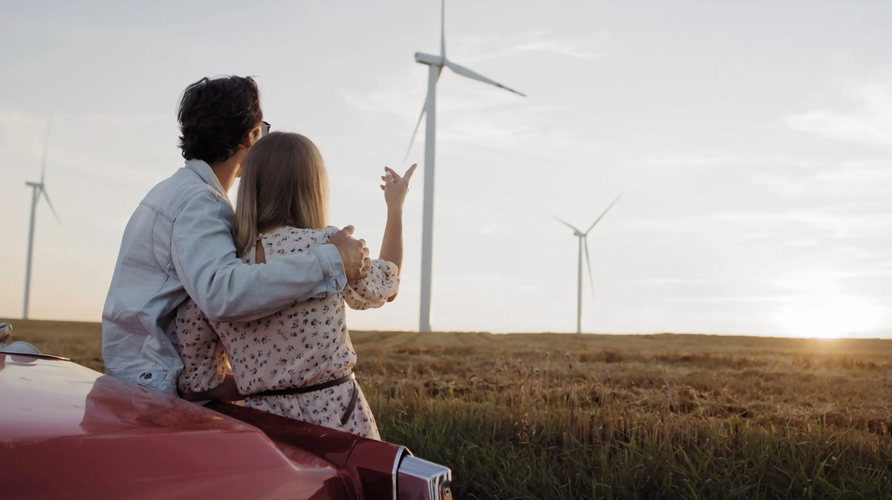 Couple sitting on the back of a red car watching wind turbines in a field at sunset.
