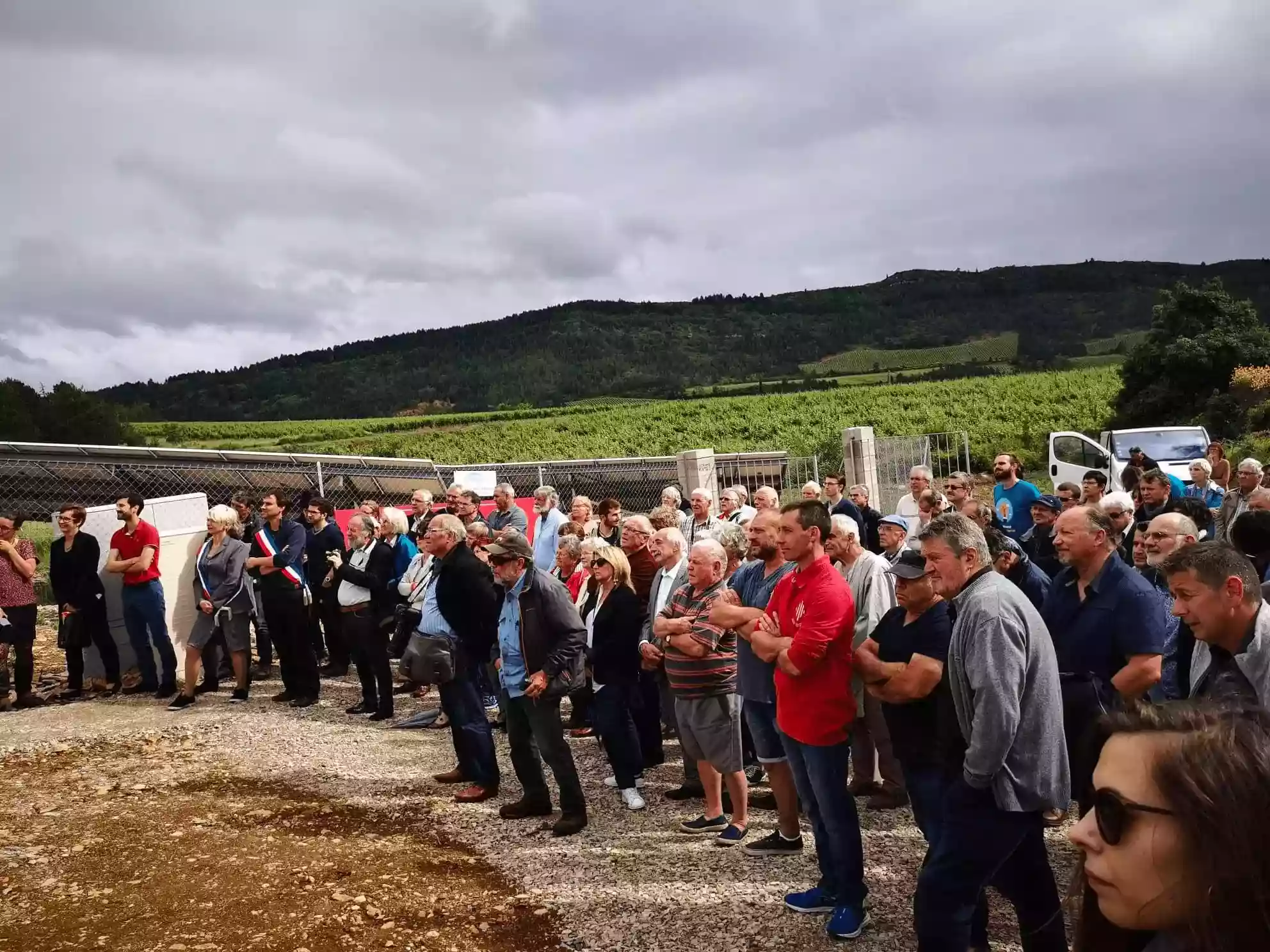 Large group of people standing outdoors on gravel near a fenced green hillside under a cloudy sky.