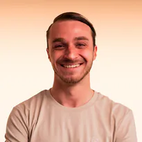Smiling young man with short hair wearing a beige t-shirt against a light background.