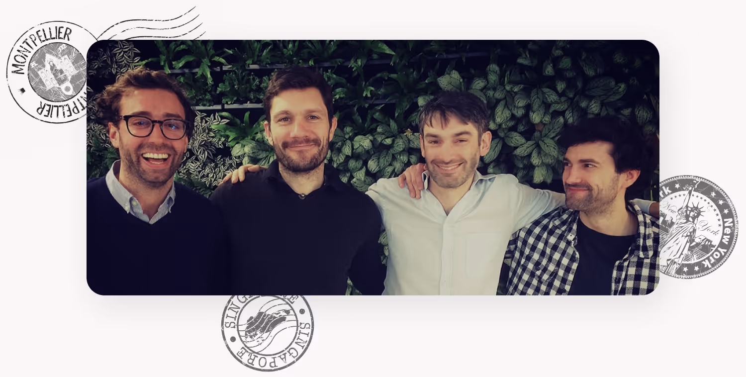 Four men standing close together in front of a leafy green wall with arms around each other, smiling.