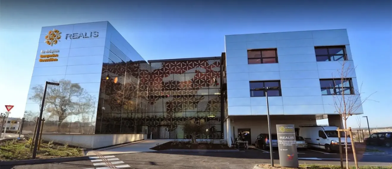 Modern office building with reflective glass facade displaying the REALIS logo and signage at the entrance under a clear blue sky.