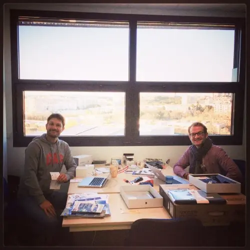 Two men sitting at a table with laptops and office supplies in front of large windows with a city view.