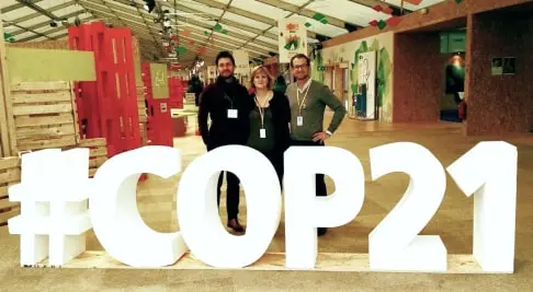 Three people standing indoors behind large illuminated white letters spelling #COP21.