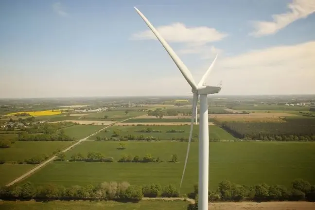 A large white wind turbine standing in a green agricultural landscape under a partly cloudy sky.