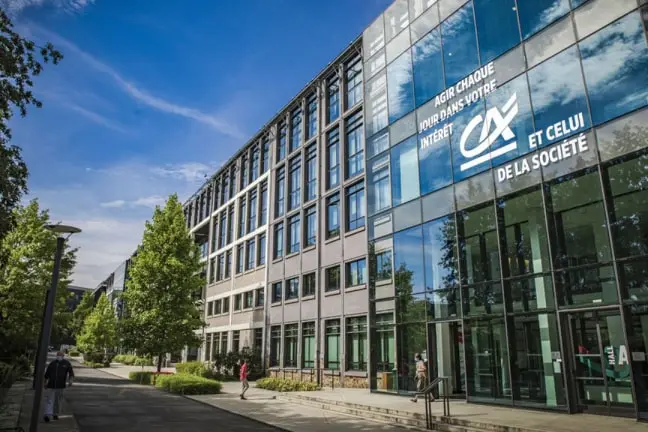 Modern office building with glass facade reflecting the sky and trees, with people walking on the sidewalk nearby.