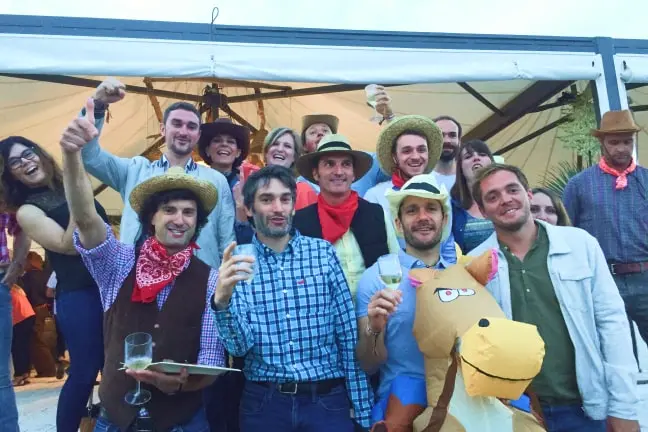 Group of people in cowboy hats and bandanas celebrating under a tent, some holding drinks and one in a horse costume.