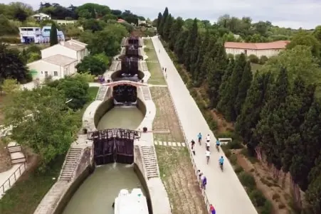 Aerial view of a canal lock system with boats and people walking along a tree-lined path beside it.