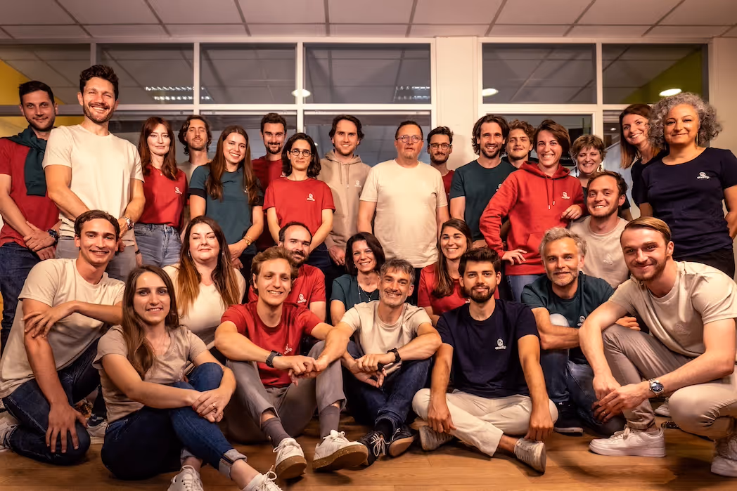 Group photo of 26 diverse people smiling and posing indoors in casual clothing.