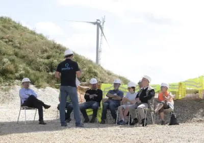 Group of people wearing hard hats seated and standing outdoors near a wind turbine and hillside.
