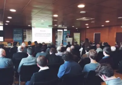 Audience attending a presentation in a conference room with a speaker at a podium and slides projected on a screen.