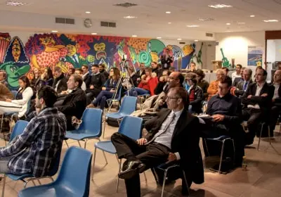 Audience seated in rows of blue chairs attentively listening in an indoor event with colorful mural art on the wall.