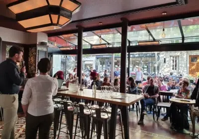 Interior of a café with people sitting at tables and two people standing near a long table with stools.