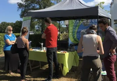 Five people interacting around an outdoor booth with a green tablecloth under a black canopy on a sunny day.