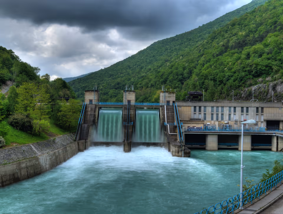 Hydroelectric dam releasing water, surrounded by green forested hills under a cloudy sky.