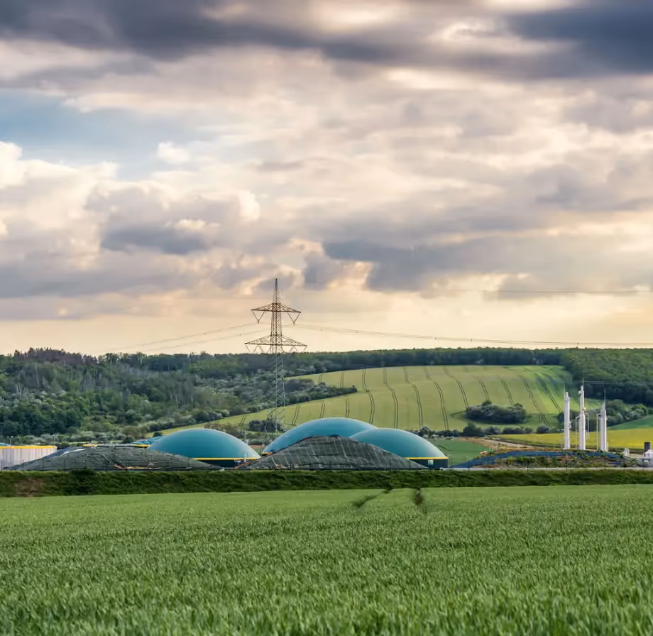 Green fields with a biogas plant featuring dome-shaped tanks and power lines under a cloudy sky.