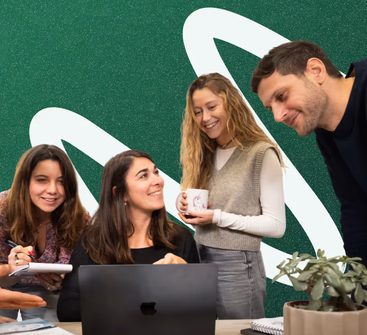 Four coworkers smiling and collaborating around a laptop at a desk with a potted plant and notebook.