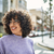 Smiling woman with curly hair wearing a light gray sweater outdoors on a sunny day.