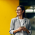 Smiling woman wearing glasses and a grey blouse standing against a yellow wall.
