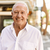 Smiling elderly man in a white shirt standing outdoors with greenery in the background.