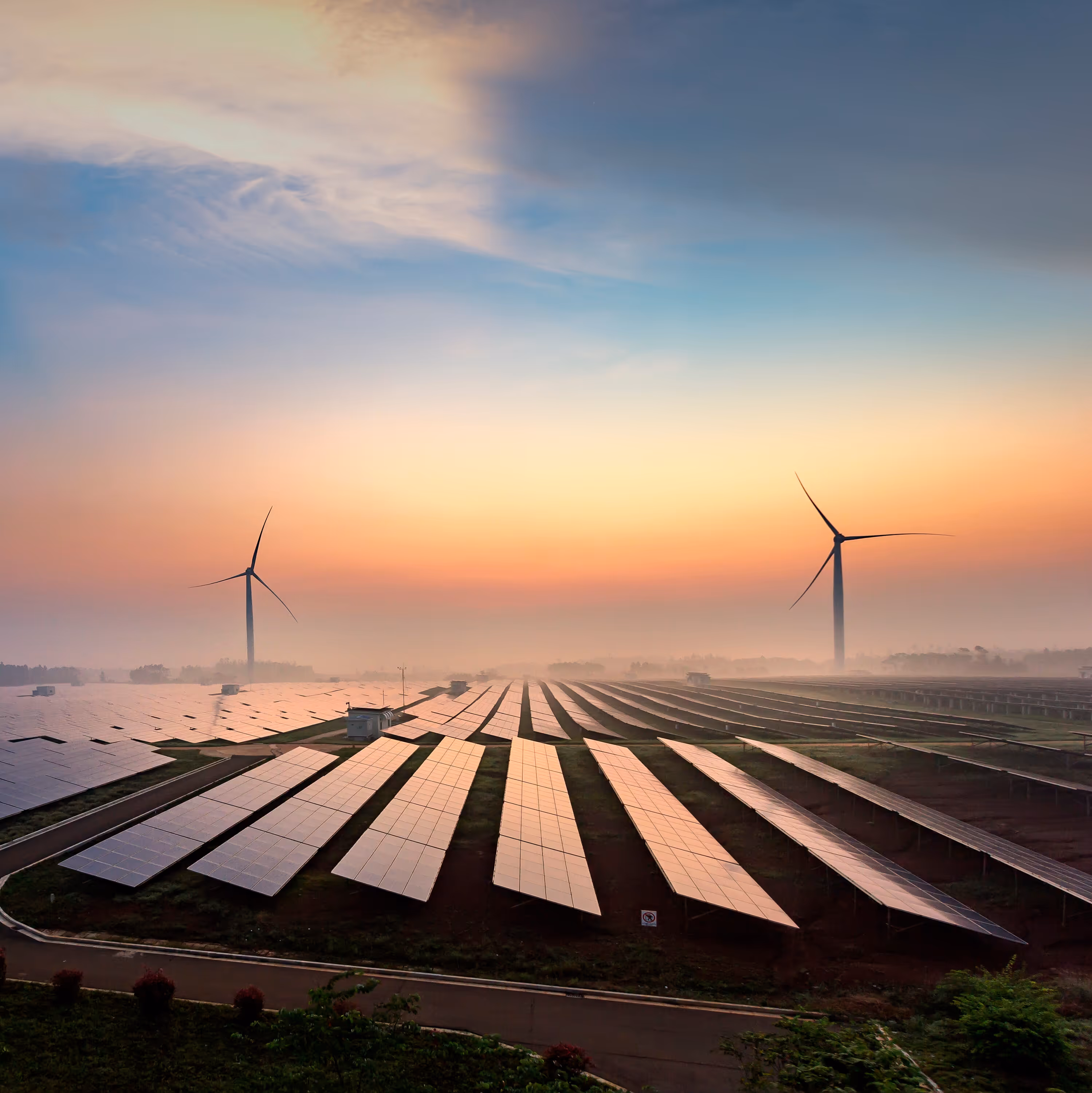 Solar panels aligned in rows with two wind turbines in the background at sunrise.
