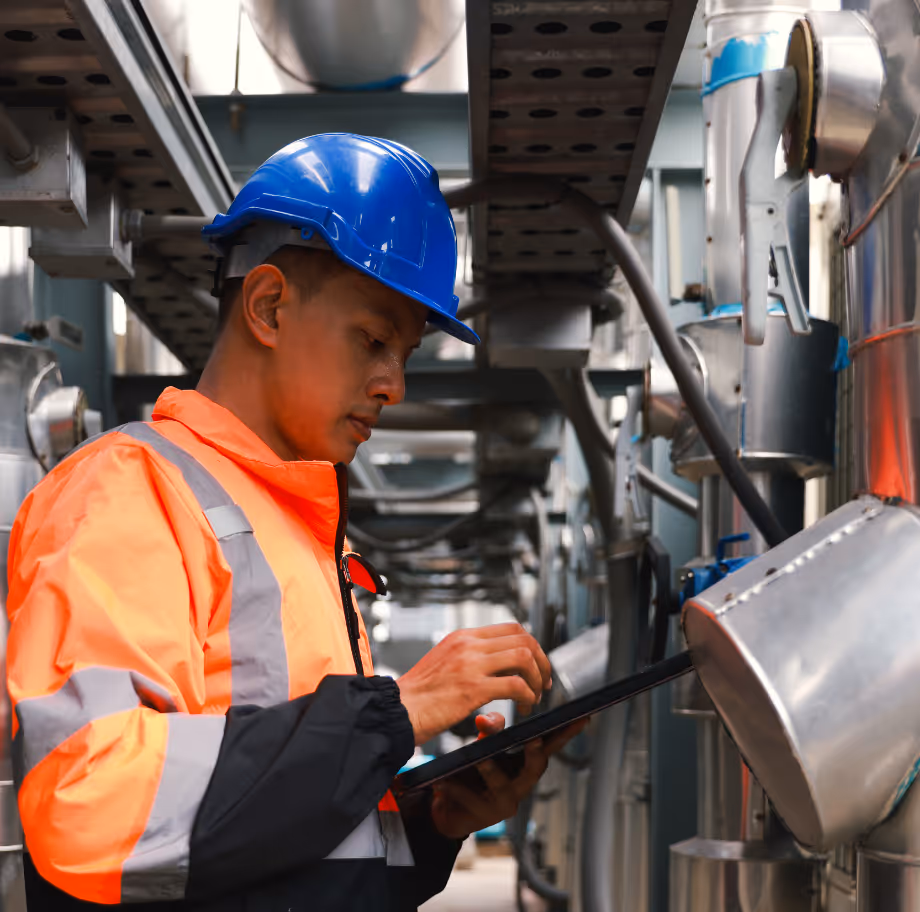 Industrial worker wearing blue hard hat and orange safety jacket using a tablet in a factory.