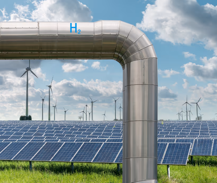 Metal pipeline labeled H2 in front of a solar panel field and wind turbines under a partly cloudy sky.