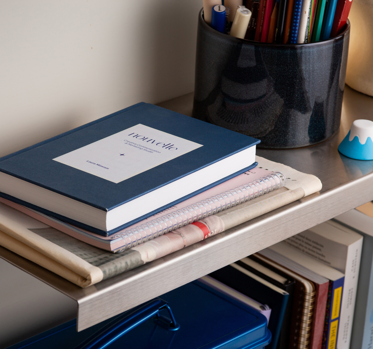 Blue hardcover book titled 'nouvelle' for Nouvelle studio in Taïwan, stacked on notebooks and newspapers on a metal shelf next to a black container holding pens and pencils.