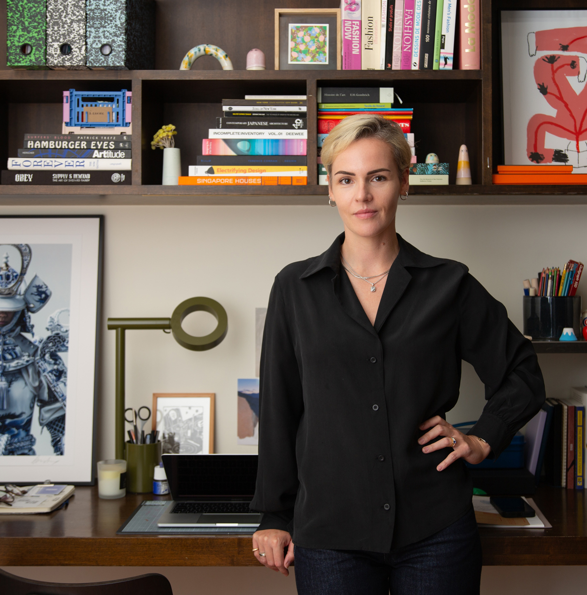 Laure Mimram with short blonde hair wearing a black shirt standing in front of a desk with a laptop and shelves filled with books and decorations.