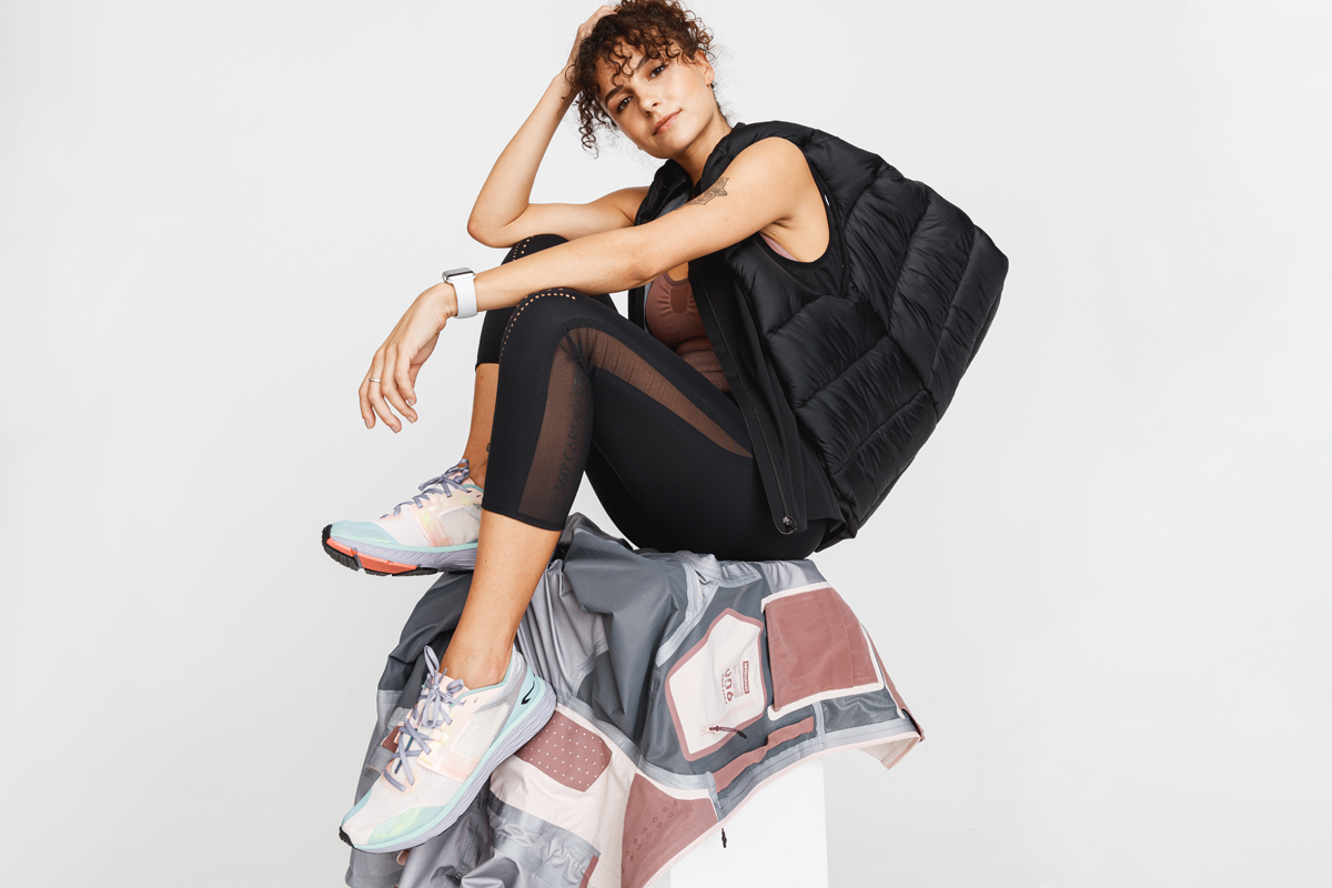 Woman wearing black puffer vest and leggings sitting on a white block draped with a gray and pink jacket. Decathlon Style Sportswear Athleisure Laure Mimram