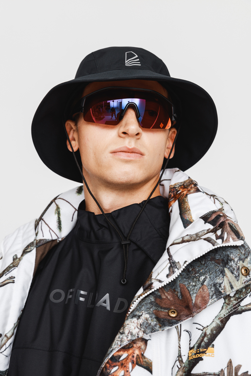 Young man wearing a black bucket hat, reflective red sunglasses, a black shirt with OFFLOAD text, and a white camouflage jacket with leaf patterns. Decathlon Style Sportswear Athleisure Laure Mimram