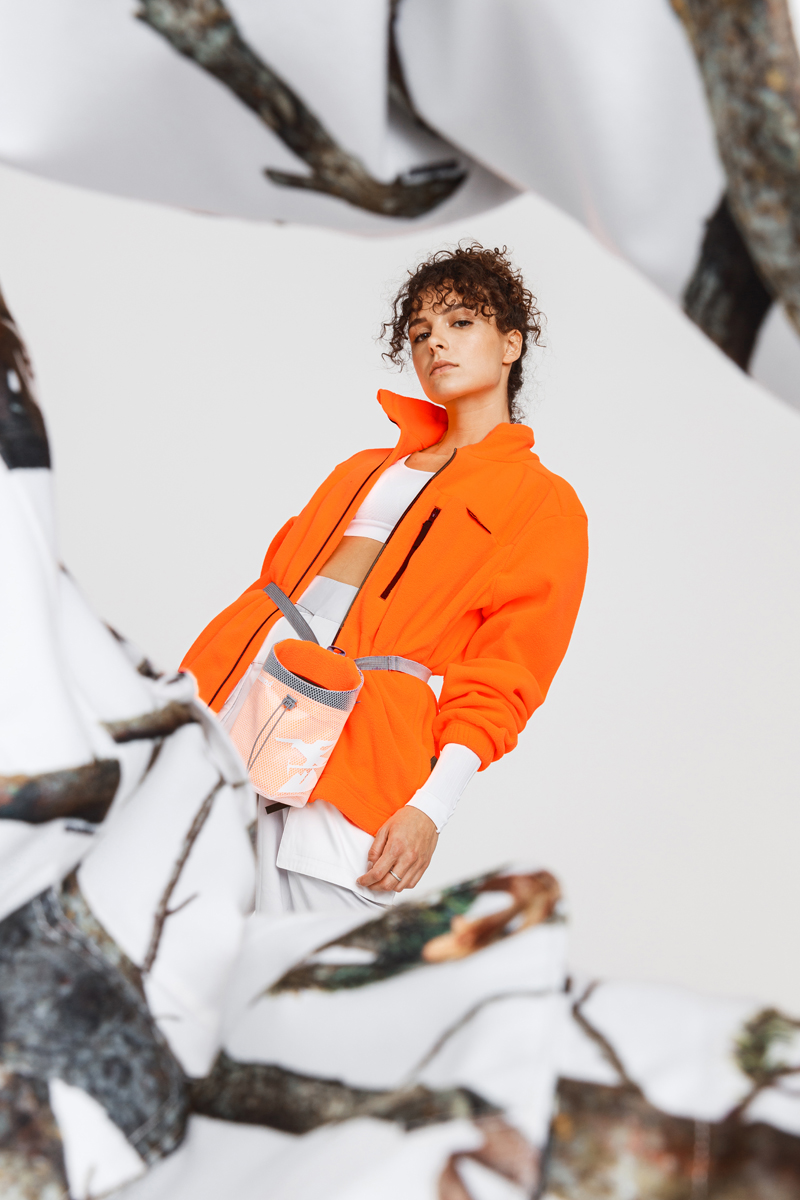 Curly-haired woman wearing a bright orange jacket and white outfit, posing with a white and orange bag against a plain background. Decathlon Style Sportswear Athleisure Laure Mimram