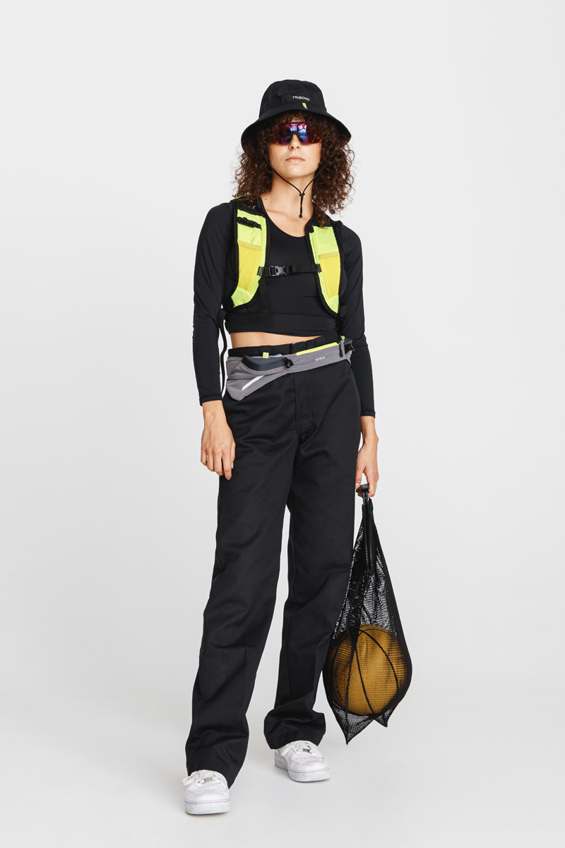 Woman in black bucket hat, sunglasses, cropped black top, and high-waisted black pants holding a black mesh bag with a basketball. Decathlon Style Sportswear Athleisure Laure Mimram