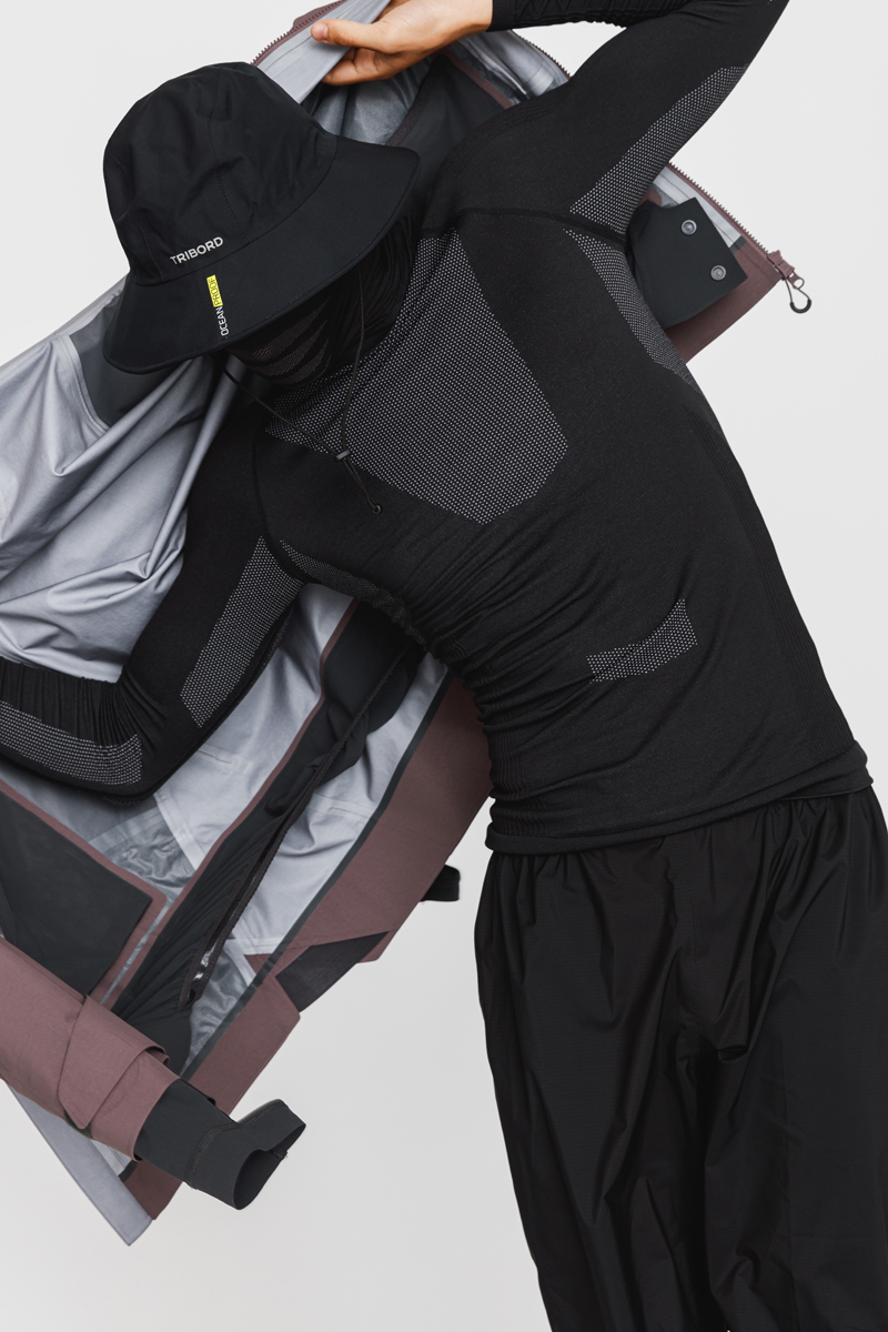 Person wearing a black patterned long-sleeve top, black pants, and a black bucket hat with 'TRIBORD' label, holding open a two-tone jacket. Decathlon Style Sportswear Athleisure Laure Mimram