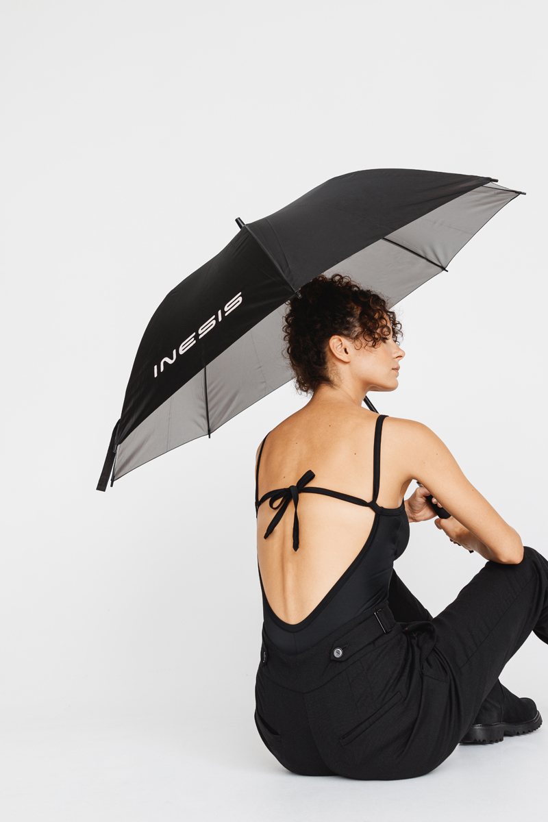 Woman sitting on the floor holding a black umbrella with 'INESIS' logo, wearing a black backless top and black pants. Decathlon Style Sportswear Athleisure Laure Mimram